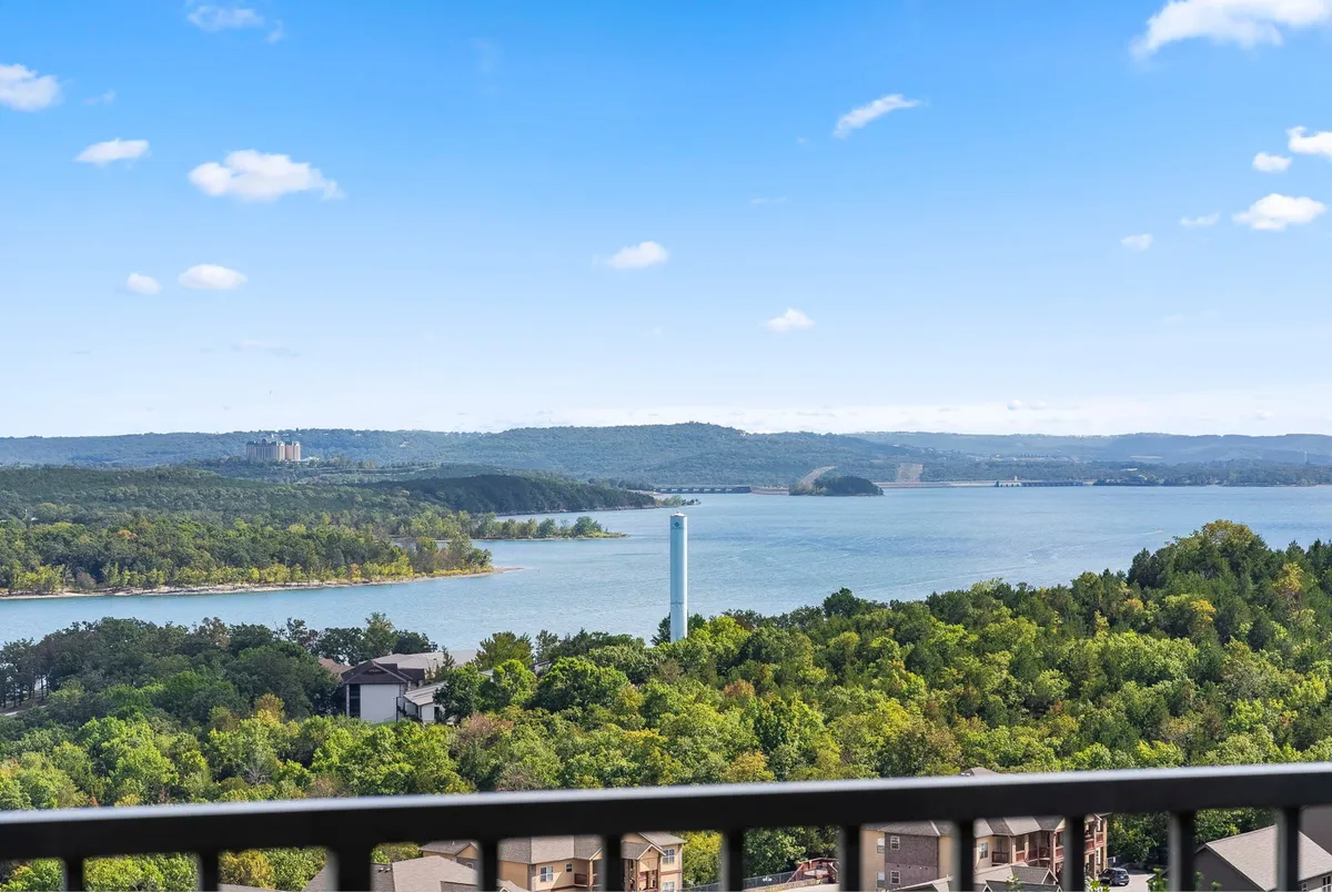 A sweeping view of Table Rock Lake bordered by lush trees and rolling hills, with buildings nestled at the water's edge and a clear blue sky overhead. The calm lake surface beckons with the promise of boating, fishing, and lakeside relaxation.