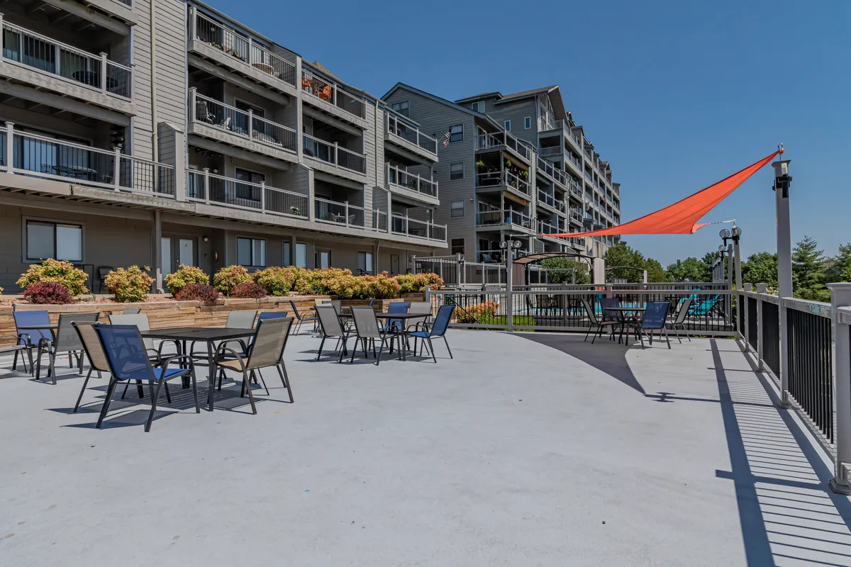 A lively exterior scene featuring an outdoor seating area under a colorful awning, surrounded by flower beds and the building façade. The setup invites group dining, social gatherings, and community events, all in a cheerful, welcoming setting.