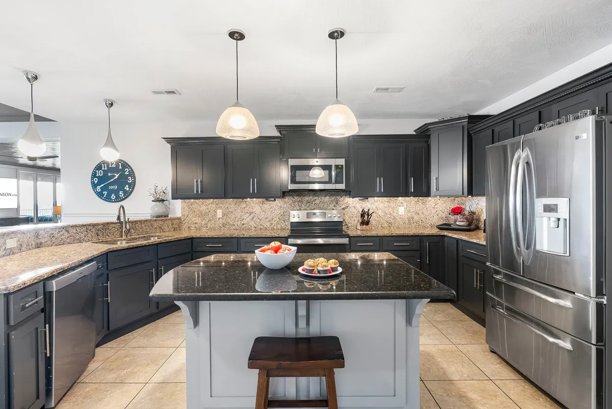 This image showcases a large kitchen island with stainless steel appliances, granite countertops, pendant lighting, and a decorative bowl of fruit. The setting is modern and open, highlighting both functionality and warmth. Bar seating and prep space are visible, indicating a kitchen designed for both meal preparation and socializing.
