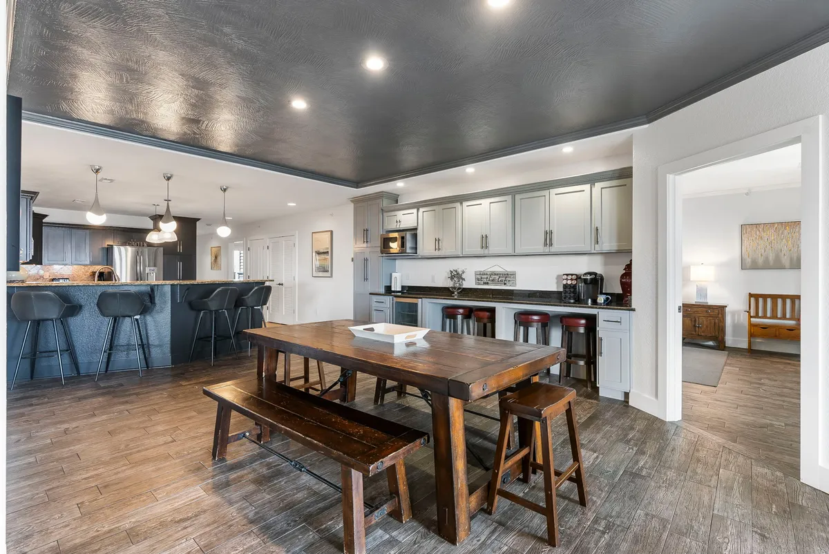 A warm and welcoming interior view featuring a large wooden dining table set near modern kitchen appliances, with bar seating and decorative wall art in the background. The space is clearly designed for family meals, cooking together, and social gatherings.