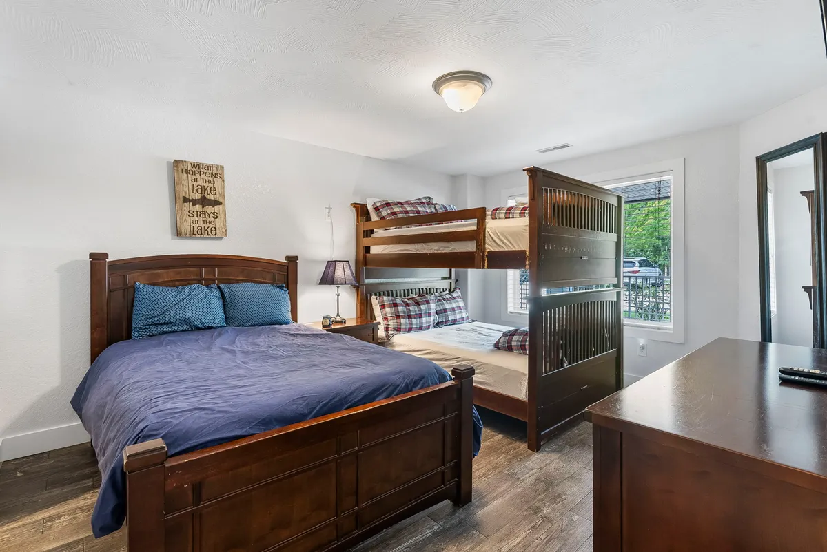 A kid-friendly bedroom featuring a bunk bed and a double bed, a nightstand with a lamp, and a window letting in daylight. The space is decorated for warmth and togetherness, suitable for families or groups with children.