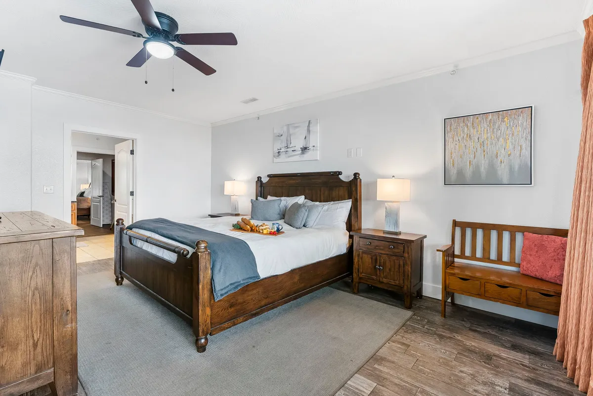 A relaxing bedroom with a king-sized wooden bed, two bedside lamps casting a warm glow, decorative artwork above the headboard, and a wooden bench at the foot of the bed. Natural light filters through unseen windows, creating a peaceful ambiance.