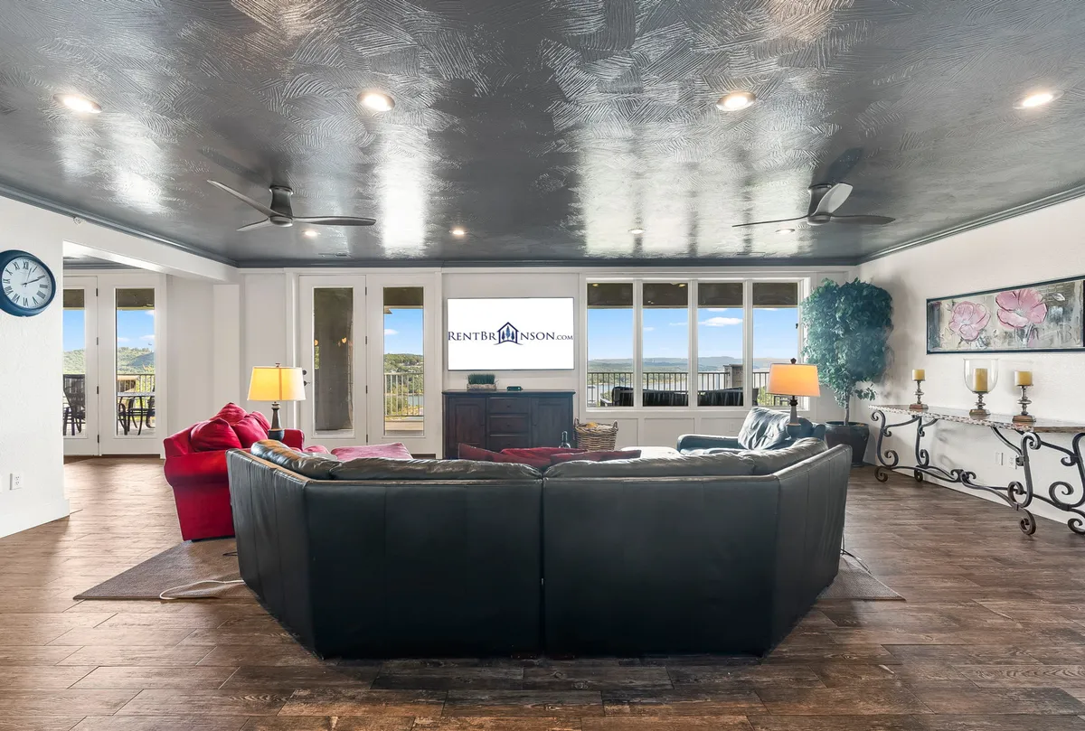 A spacious living area featuring a black sectional sofa and a red accent chair positioned near large windows that provide beautiful views and fill the room with natural light. The TV screen is visible, along with decorative wall art that adds to the cozy ambiance.