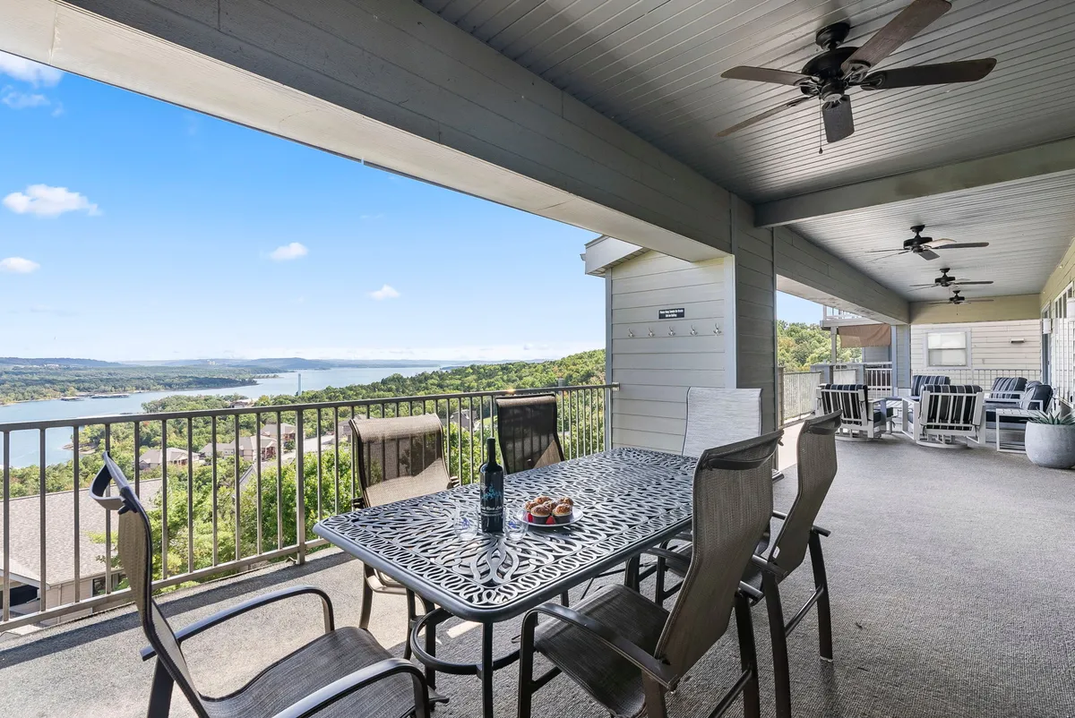 The image showcases an elegant outdoor dining area on a spacious deck overlooking a tranquil lake. A well-set dining table with comfortable chairs is positioned to take in the stunning water views, with a bottle of wine and snacks inviting guests to relax and savor the moment. The sun is setting, casting a warm glow that creates a perfect ambiance for gathering with family and friends.