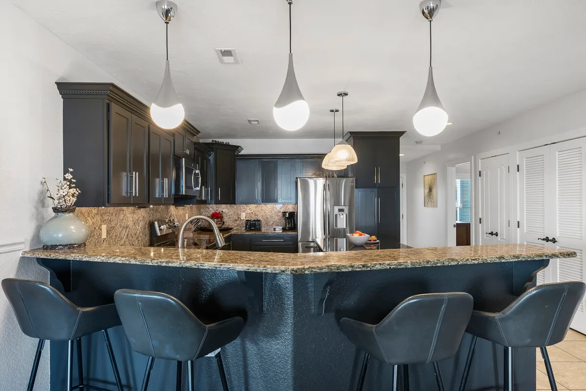 A contemporary kitchen scene featuring a granite countertop, black cabinetry, pendant lighting, and comfortable bar stools. The setting looks inviting for casual dining and group interactions, with ample space for food prep and conversation.